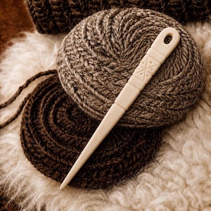 May include: A cream-coloured bone knitting needle with a geometric pattern lies on a ball of grey yarn. A second ball of dark brown yarn and knitted fabric are also visible, suggesting a crafting theme. The scene is warm and inviting.