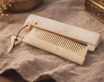 May include: A cream-colored folding comb with a smooth, polished finish and intricate circular engravings along the handle and spine. The comb features a small, triangular-shaped charm attached by a thin black cord to one end of the frame.