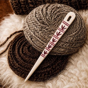 May include: A long, flat, cream-colored needle with a rounded top and a single eyelet. The needle features a series of repeating dark red leaf-like carvings along its center. It is positioned diagonally across two circular, textured yarn coils.
