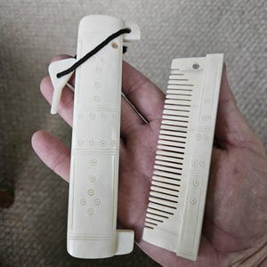 May include: Ivory-colored comb and case set. The comb has fine teeth and is rectangular. The case is also rectangular with a decorative pattern of circles and lines. A black cord is attached to the case. The set is held in a hand.