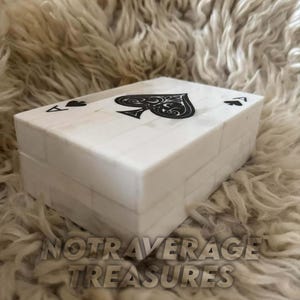 May include: A rectangular white box with a tiled surface pattern, featuring a black spade symbol centered on the lid and smaller spade symbols in the corners. The box has a smooth finish and a lid that sits flush with the base, creating a clean look.