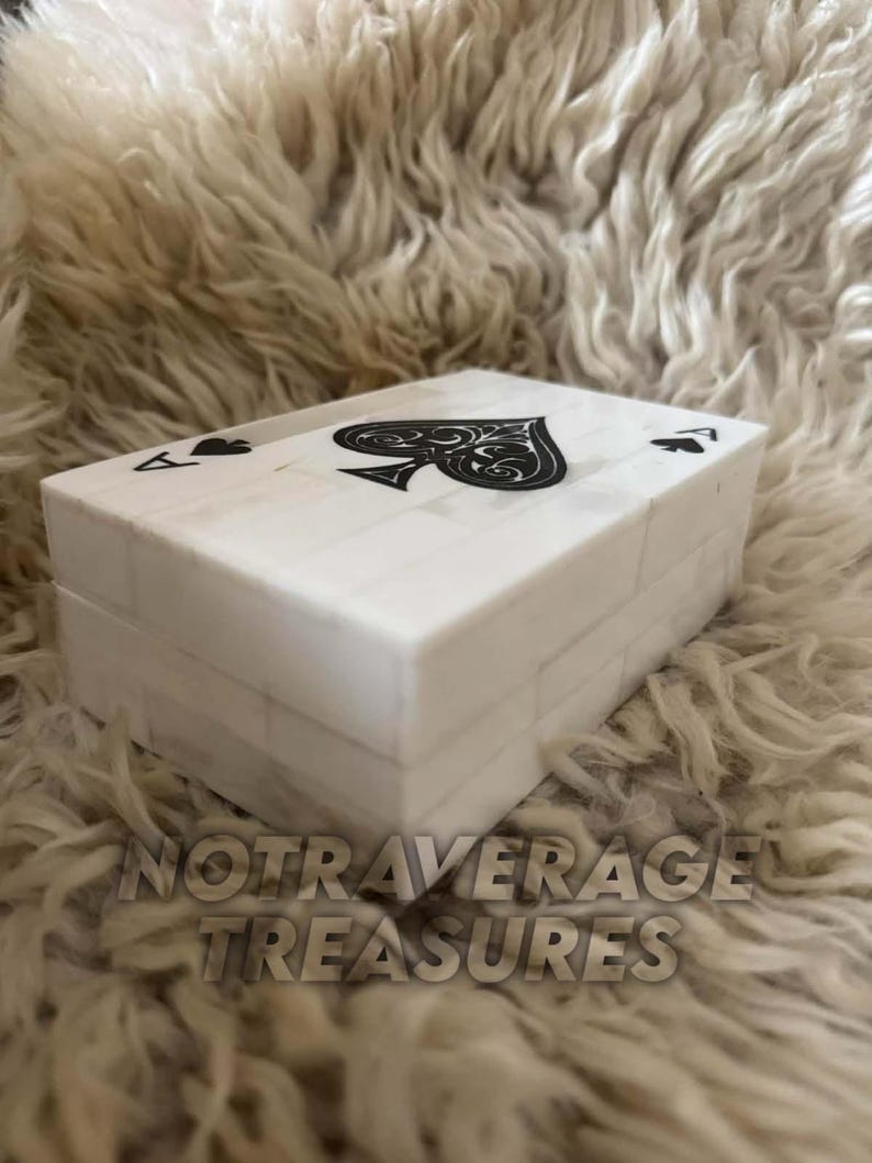 May include: A rectangular white box with a tiled surface pattern, featuring a black spade symbol centered on the lid and smaller spade symbols in the corners. The box has a smooth finish and a lid that sits flush with the base, creating a clean look.