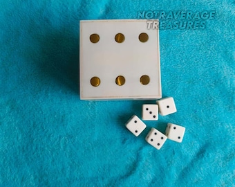 May include: A square white box with six gold circular inlays on the top surface, accompanied by five small white six-sided dice with black pips. The text NOTAVERAGE TREASURES is printed in the upper right corner of the image above the box.
