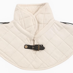 Puede incluir: Protector de cuello acolchado color crema con cierre de hebilla de cuero negro. La pieza del cuello tiene un patrón de diamantes y un cuello alto. Dos correas de cuero negro se extienden desde los lados.