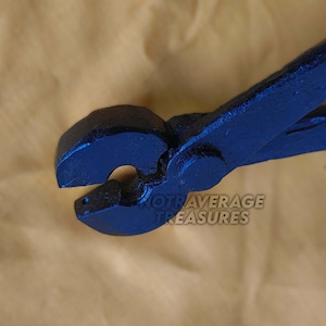 May include: Close-up of a blue metal tool with curved jaws, likely pliers or tongs. The tool has a dark blue finish and the text "NOTAVERAGE TREASURES" is visible. The background is a light brown color.
