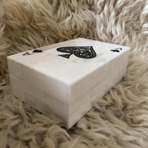 May include: A rectangular white box with a tiled surface pattern. The lid features a large black spade symbol in the center, flanked by two smaller black spade symbols and the letter A in the corners, resembling an ace of spades playing card.