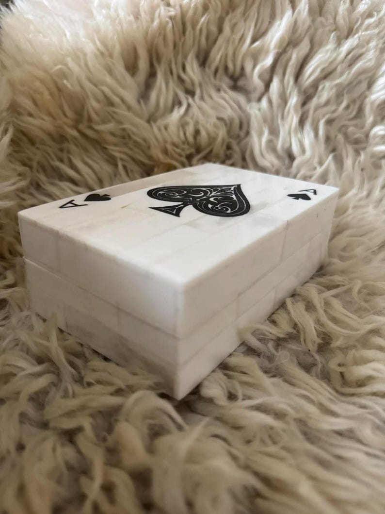 May include: A rectangular white box with a tiled surface pattern. The lid features a large black spade symbol in the center, flanked by two smaller black spade symbols and the letter A in the corners, resembling an ace of spades playing card.