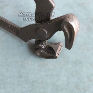 Könnte beinhalten: Eine dunkelgraue Metallzange mit texturierter Oberfläche, ausgestattet mit einem Drehpunkt und gebogenen Backen. Der Text NOTRAVERAGE TREASURES ist auf dem oberen Arm des Werkzeugs sichtbar. Die Metalloberfläche weist Gebrauchsspuren auf.