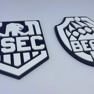 Op de afbeelding: Twee verhoogde schildvormige emblemen in zwart en wit. Het linkerembleem toont een gestileerde adelaarskop boven de tekst USEC. Het rechterembleem toont een pootafdrukontwerp boven de tekst BEAR. Beide items hebben een matte afwerking en diepte.