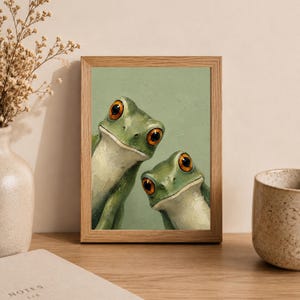 May include: A framed art print featuring two green frogs with large, orange eyes. The artwork has a light green background and is set in a natural wood frame. The frogs appear to be peeking out, creating a whimsical and playful scene.