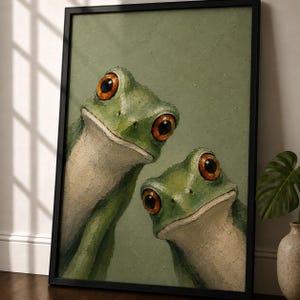 Whimsical frog wall art print featuring two expressive green tree frogs with large amber eyes painted in textured impasto oil painting style against a muted sage green background, displayed in a black frame on a wooden console table with warm sunlight, minimalist decor, ceramic vase, earthy modern interior styling, cozy cottagecore aesthetic, nature-inspired gallery wall artwork, contemporary animal painting, elegant frog poster for living room bedroom office decor