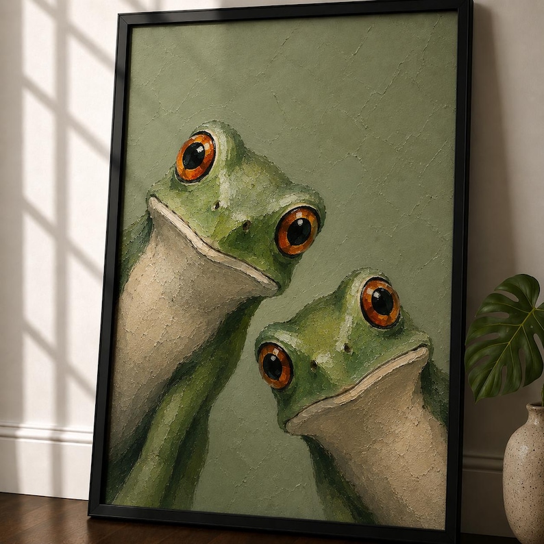 Whimsical frog wall art print featuring two expressive green tree frogs with large amber eyes painted in textured impasto oil painting style against a muted sage green background, displayed in a black frame on a wooden console table with warm sunlight, minimalist decor, ceramic vase, earthy modern interior styling, cozy cottagecore aesthetic, nature-inspired gallery wall artwork, contemporary animal painting, elegant frog poster for living room bedroom office decor