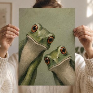 May include: A green and beige frog art print featuring two frogs with large, orange eyes. The artwork has a textured appearance and is set against a muted green background. The frogs are the focal point, with detailed facial features.