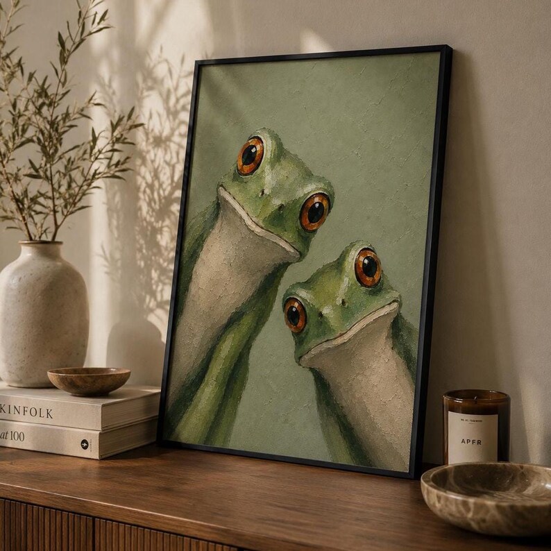 May include: A framed art print featuring two green frogs with large, orange eyes. The artwork has a light green background and is displayed in a black frame. The frogs are the focal point, creating a whimsical and nature-inspired aesthetic.