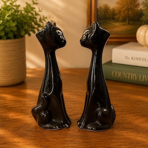 May include: Two matching black ceramic cat figurines with elongated necks and pointed ears. Each cat is depicted in a seated position with a smooth, glossy finish and stylised facial features, facing each other in a symmetrical arrangement.