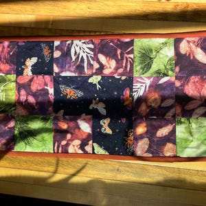 Op de afbeelding: Rechthoekige gewatteerde tafelloper met een patchwork-ontwerp. De vierkanten hebben botanische en vlinderpatronen in tinten paars, groen en rood. De loper is afgewerkt met een bruine rand.