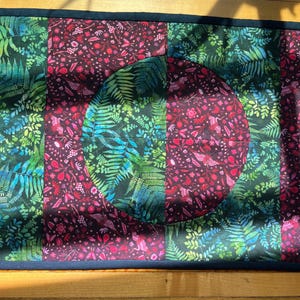 May include: A rectangular quilted table runner with a dark blue border. The runner features a patchwork design with a central circle. The fabric has a botanical print with ferns and floral elements in shades of green, red, and blue.