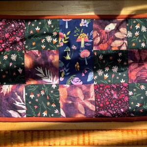Può includere: Una trapunta patchwork con una varietà di motivi floreali e botanici nei toni del bordeaux, verde e blu navy. La trapunta ha un bordo color ruggine ed è composta da più quadrati di tessuto con diversi disegni, tra cui funghi e foglie.