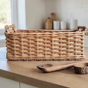 Puede incluir: Una cesta rectangular de mimbre trenzado con un acabado natural en color marrón claro. La cesta presenta un patrón de rejilla estructurado y dos asas pequeñas y resistentes en los lados izquierdo y derecho para transportar u organizar objetos.