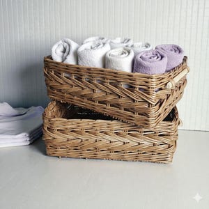 Puede incluir: Dos cestas rectangulares de mimbre trenzado apiladas una sobre otra. La cesta superior está llena de varias toallas enrolladas de color blanco y morado claro. Las cestas tienen un acabado marrón natural y asas de madera clara en los lados.