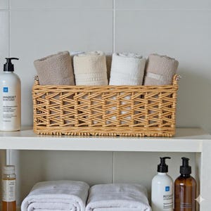 Op de afbeelding: Een geweven rieten mand gevuld met opgerolde handdoeken in beige, crème en wit staat op een witte plank. Links staat een witte fles met een zwarte pompdosator. De mand heeft een decoratief weefsel.