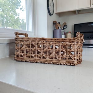 May include: A rectangular woven basket with a decorative pattern. The basket is made of light brown wicker and has a handle on each end. The basket is sitting on a white countertop.