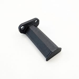 Puede incluir: Un soporte negro impreso en 3D con una base rectangular y un cuerpo hexagonal. La base tiene dos agujeros circulares. El objeto está hecho de un material texturizado y se encuentra sobre una superficie blanca.