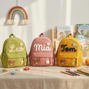 May include: Three corduroy backpacks in olive green, dusty pink, and mustard yellow. Each bag features a personalised name in white or dark blue script, a small embroidered bear face patch, and a decorative fabric flower appliqué on the front.