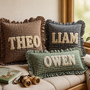 Peut inclure: Trois coussins carrés et rectangulaires avec un motif à carreaux vichy marron, noir et vert. Chaque coussin présente un bord à volants et affiche un prénom en lettres texturées en relief de couleur crème : THEO, LIAM et OWEN.