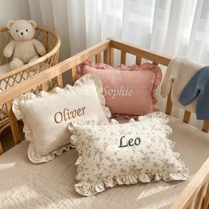 May include: A nursery setting featuring a wooden cot and a teddy bear in a wicker bassinet. Three decorative pillows with frilled edges are embroidered with the names "Oliver", "Sophie", and "Leo". Two small jumpers are also visible.
