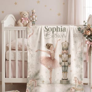 May include: A soft baby blanket featuring a printed illustration of a ballerina in a pink tutu and a nutcracker soldier. The text Sophia Brown is printed at the top. The design includes snowflakes, gift boxes, and a rocking horse in soft tones.