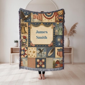 May include: A woven throw blanket featuring a patriotic patchwork design with the text James Smith in the centre. The pattern includes a bald eagle, American flag, Statue of Liberty, fireworks, a bell, a teddy bear, and a red barn with stars.