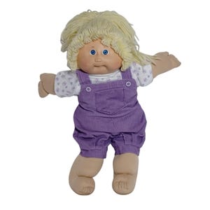 May include: A doll with light skin, blue eyes, and blonde yarn hair. The doll wears a white short-sleeved shirt with a small purple floral pattern underneath purple corduroy overalls that feature two white buttons and a front chest pocket.