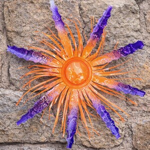 May include: A metal sunburst wall art piece with a vibrant color scheme. The center is orange, transitioning to orange and purple rays. The sunburst is mounted on a textured stone wall, creating a striking contrast.