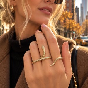 Peut inclure: Une bague dorée en forme de serpent enroulé qui entoure deux doigts. La bague présente une surface texturée rappelant des écailles sur tout le corps du serpent, avec la tête positionnée sur un doigt et la queue s'enroulant autour de l'autre.