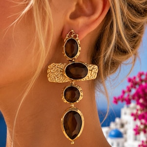 May include: A gold-toned dangle earring featuring a vertical arrangement of three dark brown oval and teardrop-shaped faceted stones. A textured, hammered gold horizontal bar sits between the top and middle stones, creating a cross-like structure.