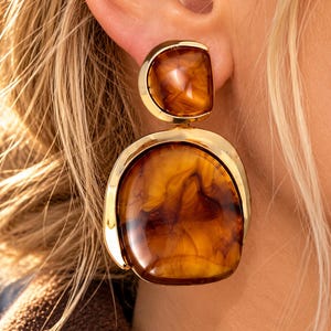 May include: A gold-toned drop earring featuring two stacked, smooth, amber-colored resin pieces with a swirling, marbled pattern. The top piece is a smaller semi-circle, while the bottom piece is a larger, rounded, irregular shape.