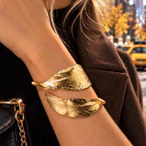May include: A gold-toned cuff bracelet featuring two overlapping leaf-shaped elements with a textured, organic surface. The metal has a polished, reflective finish that catches the light, creating a distinct contrast against the dark clothing.