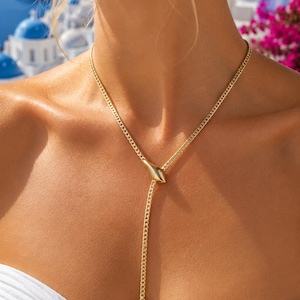 Op de afbeelding: Een goudkleurige lariat-ketting met een fijne gourmetketting. Het ontwerp bevat een glad, druppelvormig centraal verbindingsstuk en een enkele, puntige conische hanger die aan het uiteinde van de ketting hangt en op de borst rust.