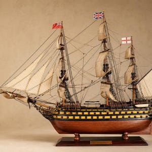May include: A detailed wooden model of a sailing ship, with multiple masts and sails, and various flags. The ship has a dark brown hull with yellow and black accents, and is mounted on a wooden base. The sails are off-white.