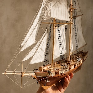 May include: A finely crafted wooden model of a sailing ship, showcasing white sails and complex rigging. The ship has a brown hull and deck, with detailed masts, ropes, and anchors. The sails are partially open, giving the impression of a vessel at sea.