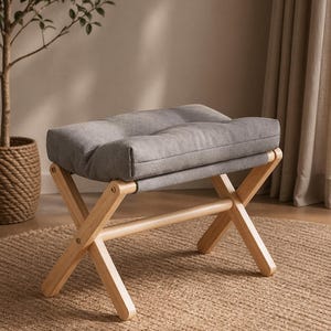 May include: A grey upholstered ottoman with a tufted top on a natural wood X-frame base. The ottoman is on a woven jute rug. A small potted tree is in the background.