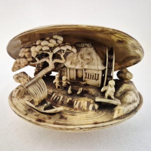 May include: An off-white, carved clam shell sculpture featuring a detailed miniature scene of a traditional house, a water wheel, a small boat, and figures. The interior of the shell is intricately sculpted with landscape elements and structures.