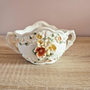 May include: Cream ceramic planter with a scalloped rim and two handles. The planter features raised floral designs in yellow, orange, and green. The planter is on a wooden surface.