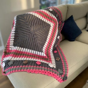May include: A handmade crochet blanket in shades of gray, white, pink, and brown, draped over a beige sofa. The blanket features a square central design, bordered by stripes and adorned with flower appliqués. The edges are finished with a scalloped pink trim.