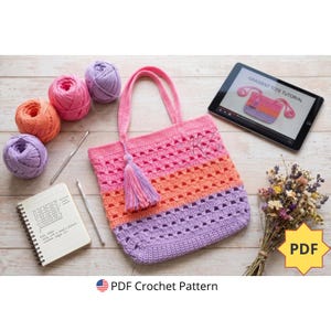 Peut inclure: Un sac fourre-tout crocheté aux tons rose, orange et violet, avec un pompon assorti. L'image présente des pelotes de laine, des crochets, un carnet avec des notes de motifs et une tablette affichant un tutoriel. Le texte "PDF Crochet Pattern" est également visible.