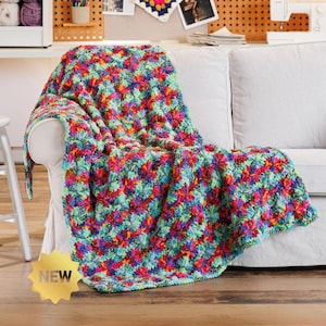 Peut inclure: Une couverture en crochet multicolore présentant un motif texturé et vibrant de fils entrelacés rouges, bleus, verts, jaunes et violets. La couverture est drapée sur un canapé clair et comporte un badge doré en forme d'étoile avec le texte NEW.