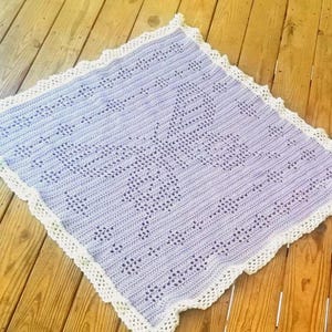 Peut inclure: Une couverture au crochet violet clair faite à la main avec un bord festonné blanc. La couverture présente un motif ajouré détaillé et est présentée sur une surface en bois. La couverture est de forme carrée.
