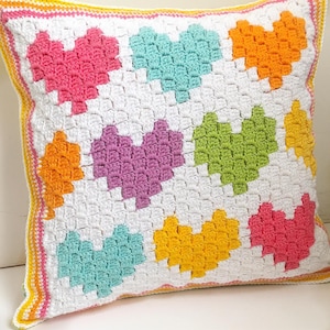 Peut inclure: Un coussin carré en crochet blanc avec un motif de cœurs colorés. Les cœurs sont dans des tons de rose, turquoise, orange, violet, vert et jaune. Le coussin a une bordure blanche avec une garniture jaune et rose.