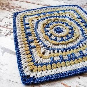 Peut inclure: Un ouvrage carré au crochet, fait à la main, avec un motif en spirale au centre. La pièce présente une palette de couleurs composée de bleu, beige et crème. La bordure est bleu foncé, et la texture est complexe et détaillée.